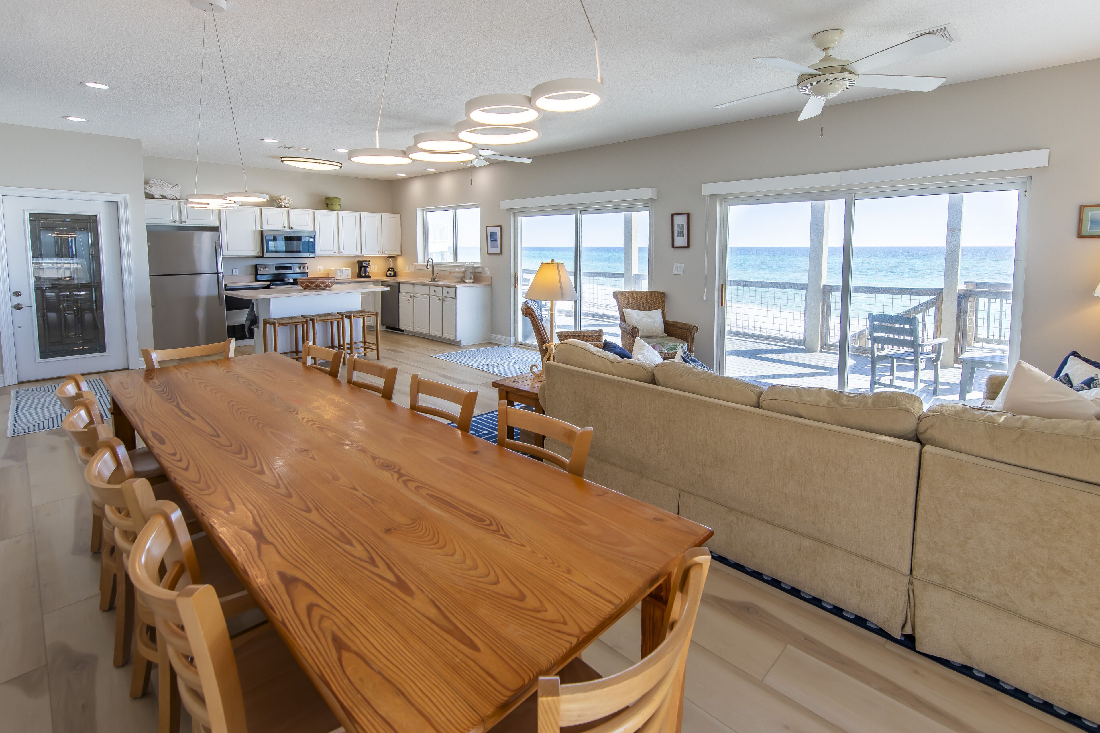 A spacious kitchen with a wooden dining table and chairs, a sofa facing a large sliding glass door to the ocean, and a ceiling fan.