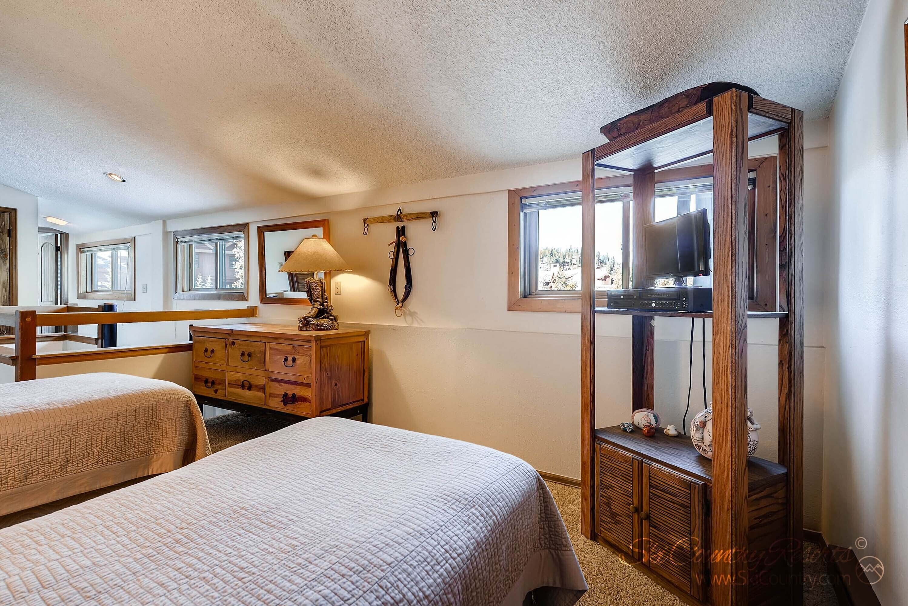 Une chambre avec deux lits, une commode en bois, un téléviseur posé sur un meuble en bois, et une fenêtre donnant sur des arbres.