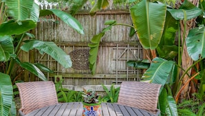 A round wooden table with two wicker chairs, surrounded by lush green foliage and a decorative wooden wall with a circular emblem. - Historic Downtown Cottage with Private/Heated Pool! Walk to everything! (st. augustine)