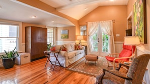 A cozy living room with a wooden floor, a sofa, a red chair, and a dining table. - Historic Downtown Cottage with Private/Heated Pool! Walk to everything! (st. augustine)