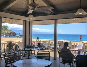 A covered outdoor seating area with tables and chairs, overlooking a beach with people and umbrellas. - Maui Eldorado B206 Spacious Corner Unit w/Premium Upgrades & Lanai (Kaanapali)