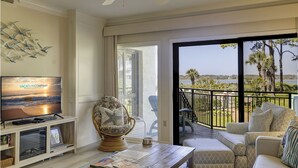 A spacious living room with a television, a comfortable armchair, a sofa, and a balcony with a view of the ocean.