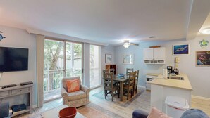A living room with a TV, a sofa, a dining table with chairs, a kitchen area with cabinets, and a balcony with a view of trees. - Villa 212 3 bedrooms, 2 bathroom (Jekyll Island)