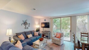 A living room with a grey sofa, a small table, and a TV on the wall. There is a balcony with a wooden table and chairs. - Villa 212 3 bedrooms, 2 bathroom (Jekyll Island)