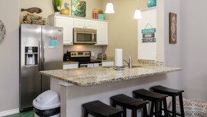 A modern kitchen with a stainless steel refrigerator, a microwave, a water dispenser, granite countertops, and a bar stool area. - Poolside Coastal Retreat | Short Walk to the Beach (Corpus Christi)