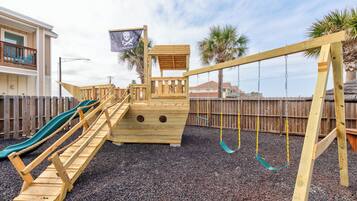 Une structure de jeu en bois comprenant un toboggan, une petite maison de jeu sur le thème des pirates et un ensemble de balançoires.