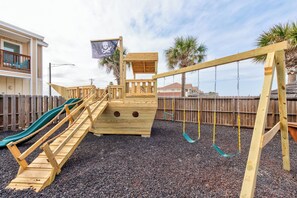 Une structure de jeu en bois comprenant un toboggan, une petite maison de jeu sur le thème des pirates et un ensemble de balançoires.