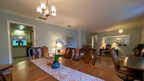Comedor con mesa de madera, sillas y candelabro. Se ve una cocina al fondo.