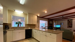 Una cocina con armarios blancos, lavavajillas y ventana con cortinas. Hay un comedor con mesa y sillas, y una sala de estar con sofá y televisor.