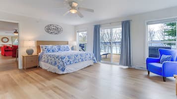 A bedroom with a large bed, a ceiling fan, a view of the outdoors, and a red sofa in the adjacent room.
