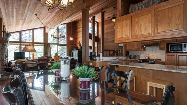 Un interior espacioso de madera con una gran mesa de comedor rodeada de sillas, una zona de cocina con armarios de madera y una chimenea de piedra.