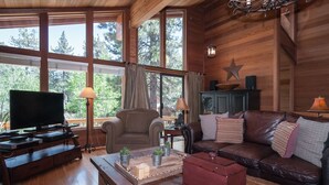 Una sala de estar acogedora con televisión, sofá de cuero, mesa de centro de madera y amplios ventanales con vistas a árboles.