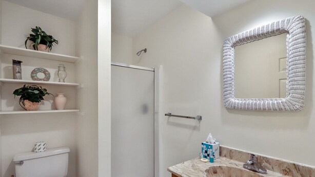 Un baño con inodoro, ducha y lavamanos. Hay estantes con objetos decorativos y un espejo sobre el lavamanos.