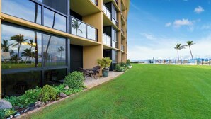 A modern hotel with a well-maintained lawn, outdoor seating area, and large windows offering views of the ocean and palm trees.