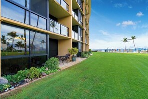 A modern hotel with a well-maintained lawn, outdoor seating area, and large windows offering views of the ocean and palm trees.