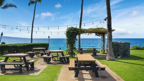An outdoor seating area with picnic tables and string lights, overlooking a body of water. - Oceanfront Ground-Floor Retreat: Kihei Surfside #111 – Stunning Views! (Kihei, Maui)