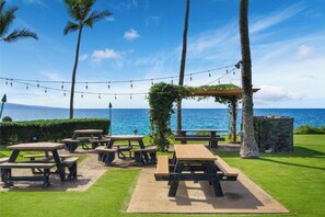 An outdoor seating area with picnic tables and string lights, overlooking a body of water. - Oceanfront Ground-Floor Retreat: Kihei Surfside #111 – Stunning Views! (Kihei, Maui)