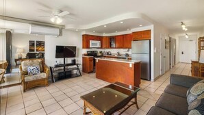 A modern kitchen with wooden cabinets, a central island, and a built-in microwave. There is a dining area with a glass table and chairs, and a living room with a sofa and a TV. - Oceanfront Ground-Floor Retreat: Kihei Surfside #111 – Stunning Views! (Kihei, Maui)