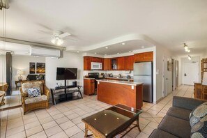 A modern kitchen with wooden cabinets, a central island, and a built-in microwave. There is a dining area with a glass table and chairs, and a living room with a sofa and a TV. - Oceanfront Ground-Floor Retreat: Kihei Surfside #111 – Stunning Views! (Kihei, Maui)