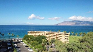 A hotel with a parking lot, surrounded by greenery and a view of the ocean. - Oceanfront Ground-Floor Retreat: Kihei Surfside #111 – Stunning Views! (Kihei, Maui)