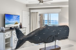 En stue med et fjernsyn der viser et strandbillede, en stor hvalskulptur og udsigt til havet gennem en skydedør.