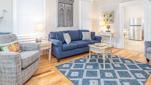 A living room with a blue sofa, a small coffee table, and a patterned rug. There is a window with blinds, a ceiling fan, and a kitchen area visible in the background. - Sea View 2 | Pet Friendly, Large Unit, Near Beach! (Tybee Island)