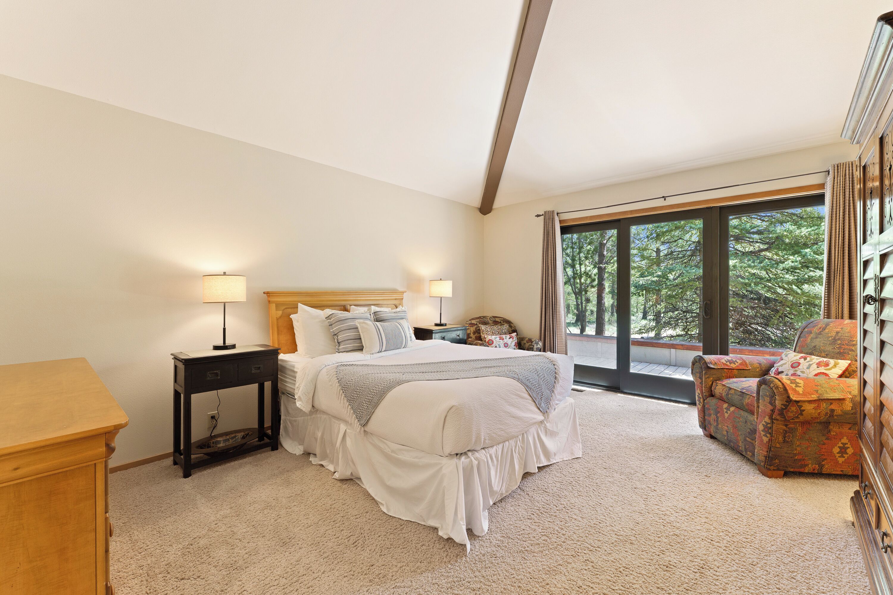 A bedroom with a large bed, bedside tables, a chair, and a view of trees through large windows.