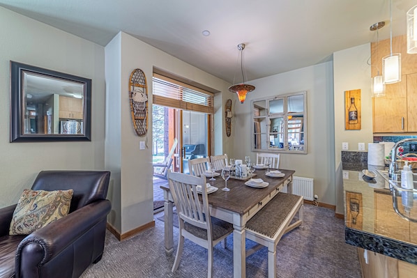 A dining area with a wooden table set for four, surrounded by white chairs. A leather sofa with a patterned throw pillow is positioned to the left. - Golf Course Townhome, Views & Spa 320 (Mammoth Lakes)