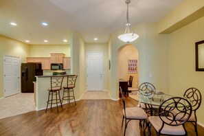 A modern kitchen with wooden cabinets, a stainless steel refrigerator, and a microwave. There is a dining area with a glass table and metal chairs.
