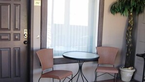 Een patio met een deur, een raam met gordijnen, twee rieten stoelen, een kleine ronde tafel en een bloempot.
