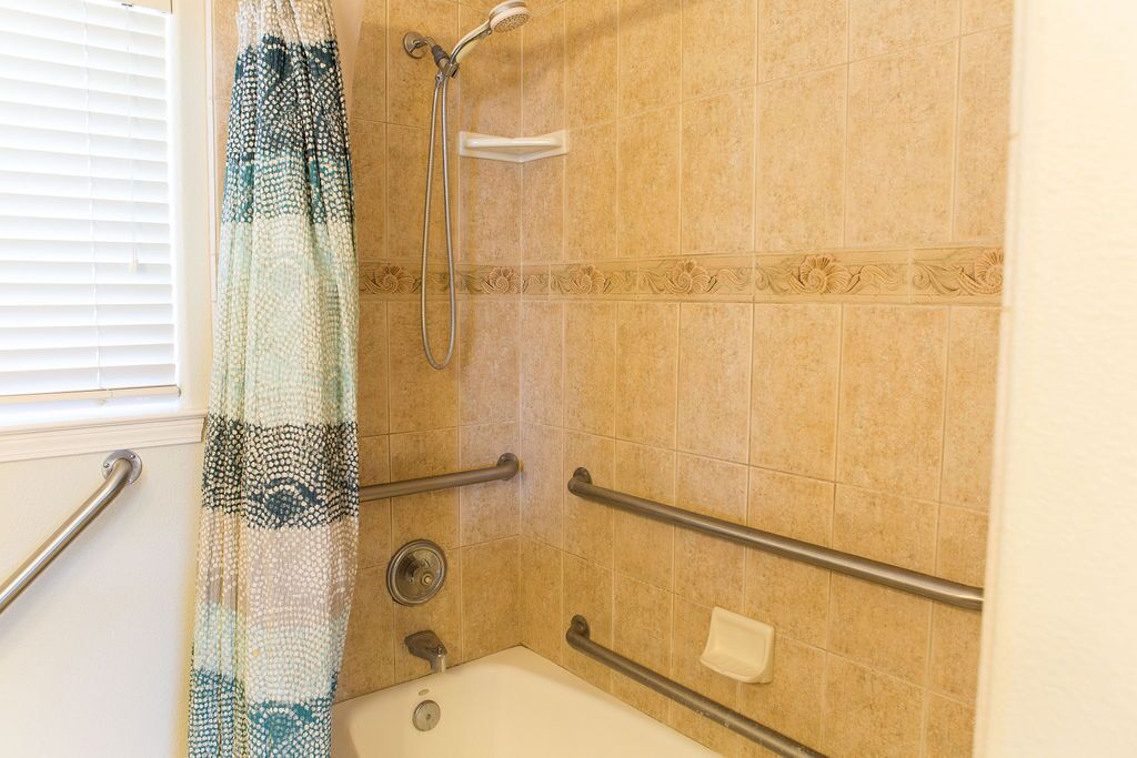 Ein Badezimmer mit beigen Fliesen, einer Dusche mit Duschbrause, einer weißen Badewanne und einem gemusterten Vorhang.