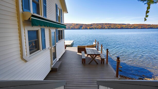 Eine Holzterrasse mit Tisch und Stühlen mit Blick auf einen See, ein Haus ist links zu sehen.