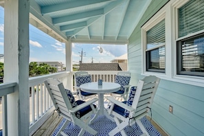 A covered porch with a white table and blue chairs, a white railing, and a view of the street and houses in the background. - Ryan's Retreat - Retreat to this charming cottage at the coveted south end (Wrightsville Beach)