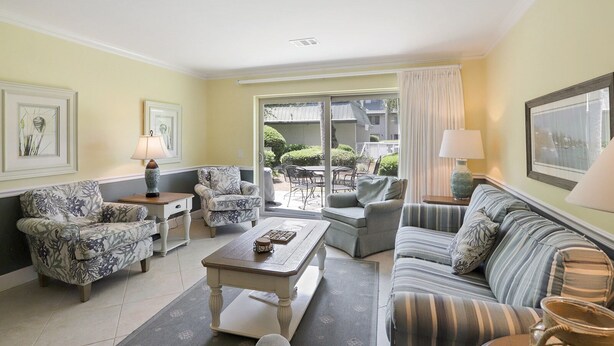 A living room with a sofa, armchairs, a coffee table, and a dining table. There is a sliding glass door leading to an outdoor patio area.