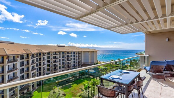A balcony with a glass railing overlooking a resort area, a dining table with chairs, and a view of the ocean and hotel buildings.