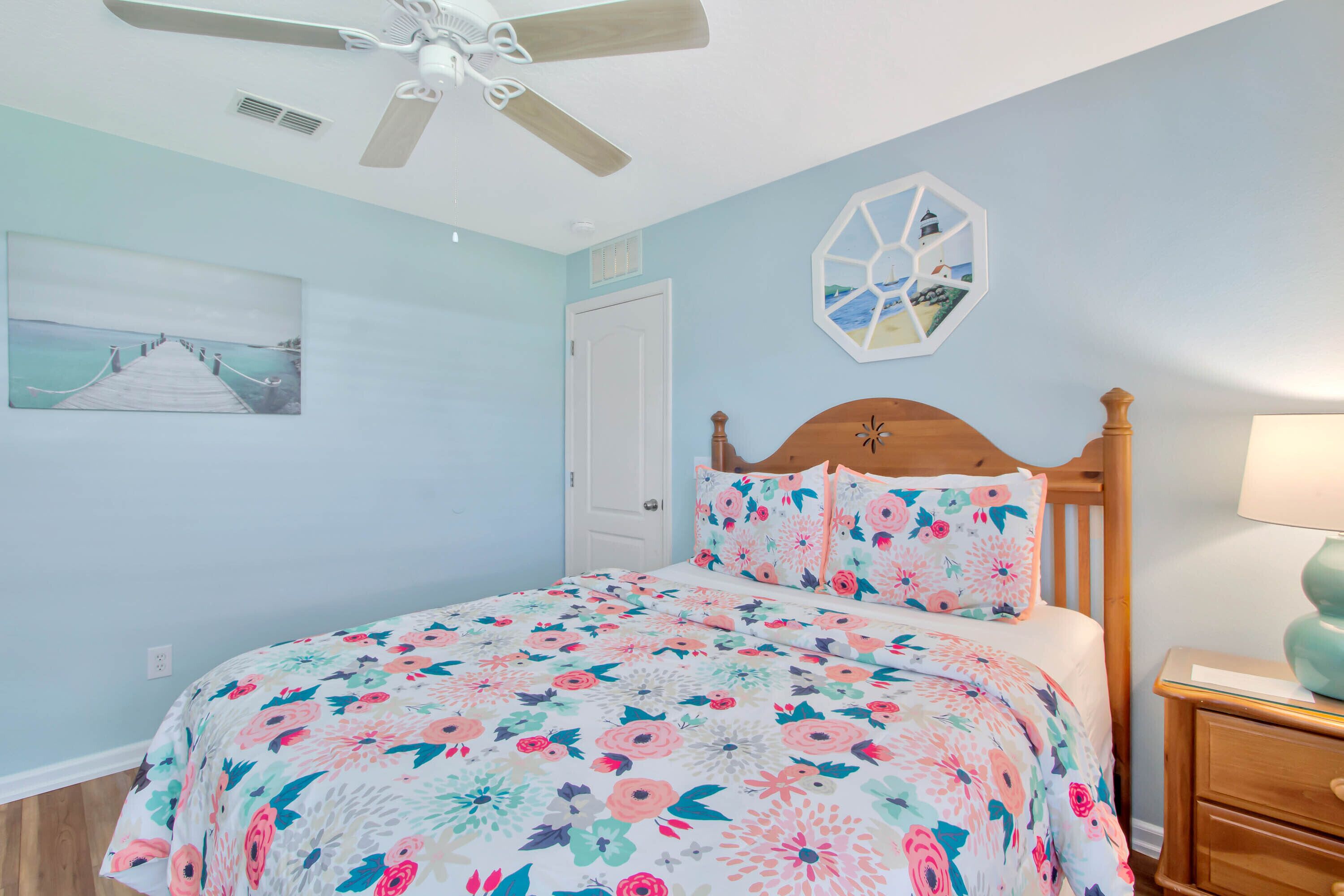 A bedroom with a floral bedspread, a wooden headboard, a ceiling fan, a wall-mounted artwork, and a nightstand with a lamp.