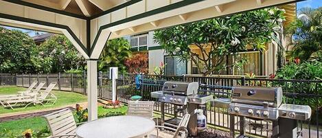 An outdoor patio with a round table and chairs, a grill, and a covered area with a view of a garden and a house.