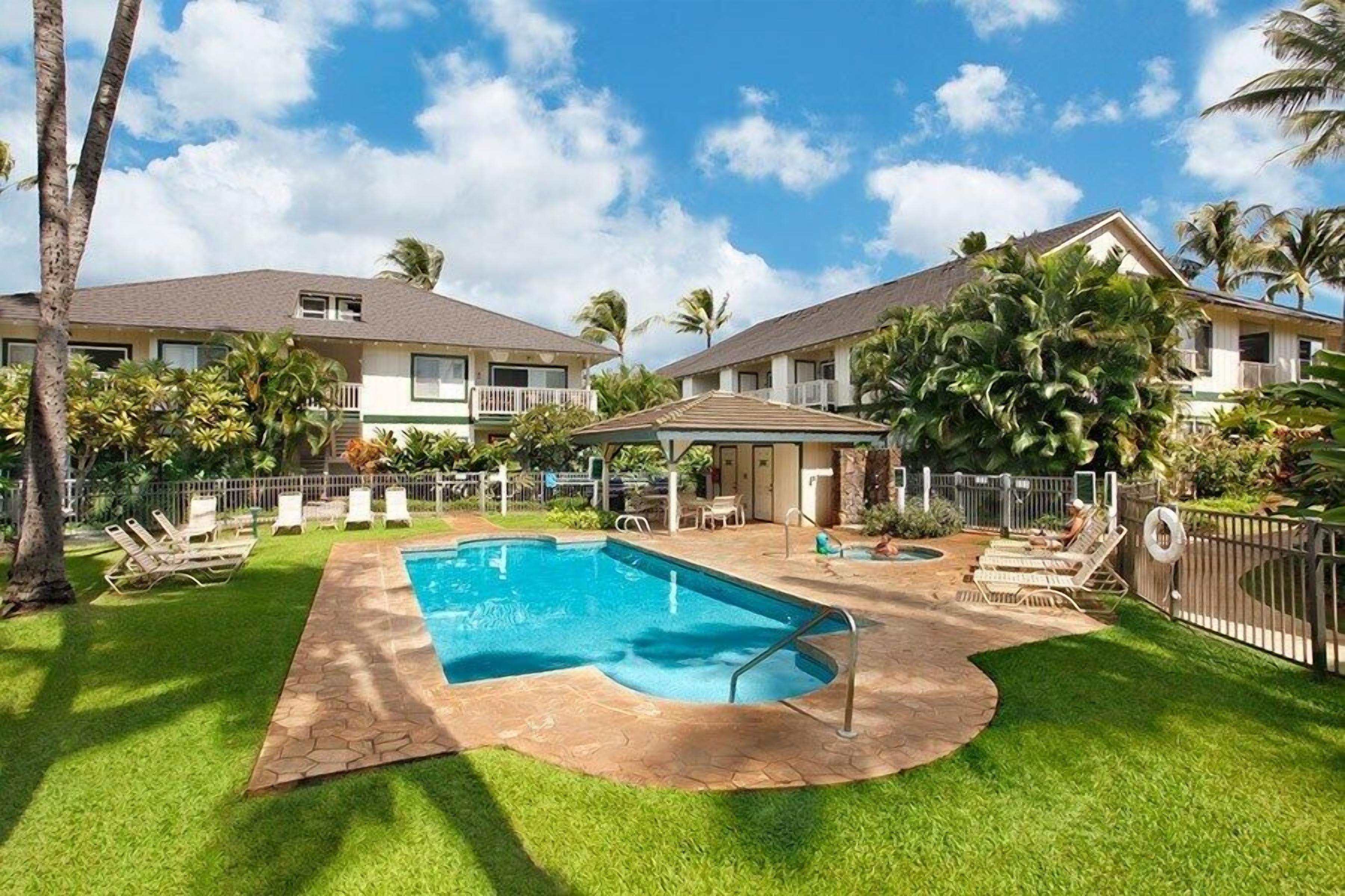 A pool area with a clear blue pool, surrounded by a stone deck and green grass. There are lounge chairs and a covered seating area.