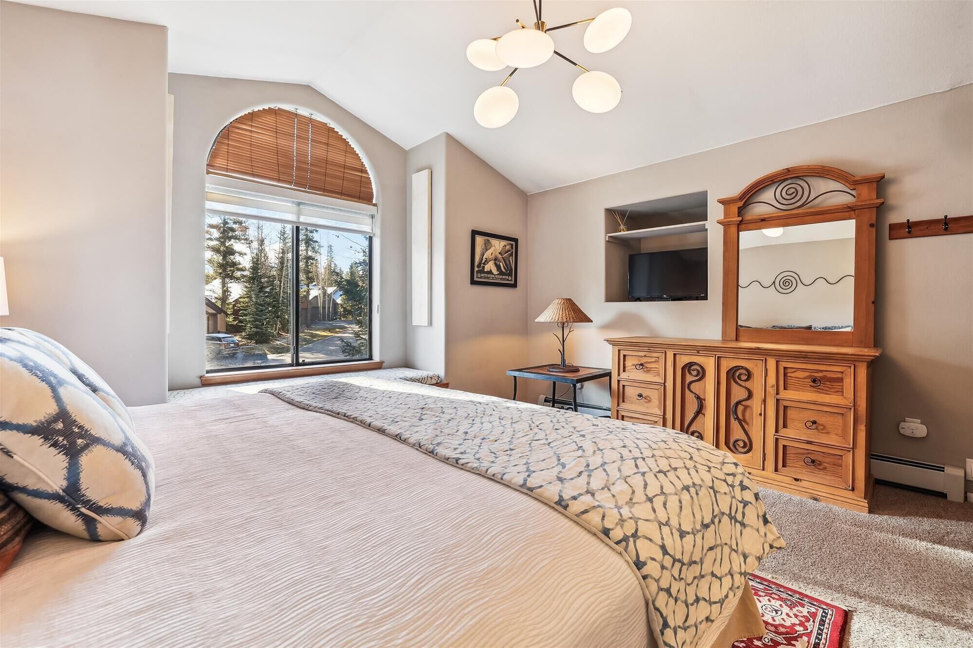 A bedroom with a large bed, a wooden dresser, a TV, and a window with blinds.