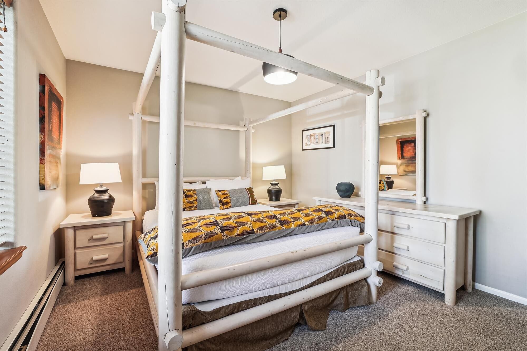 A four-poster bed with a patterned bedspread, bedside tables with lamps, a dresser, and a framed picture on the wall.