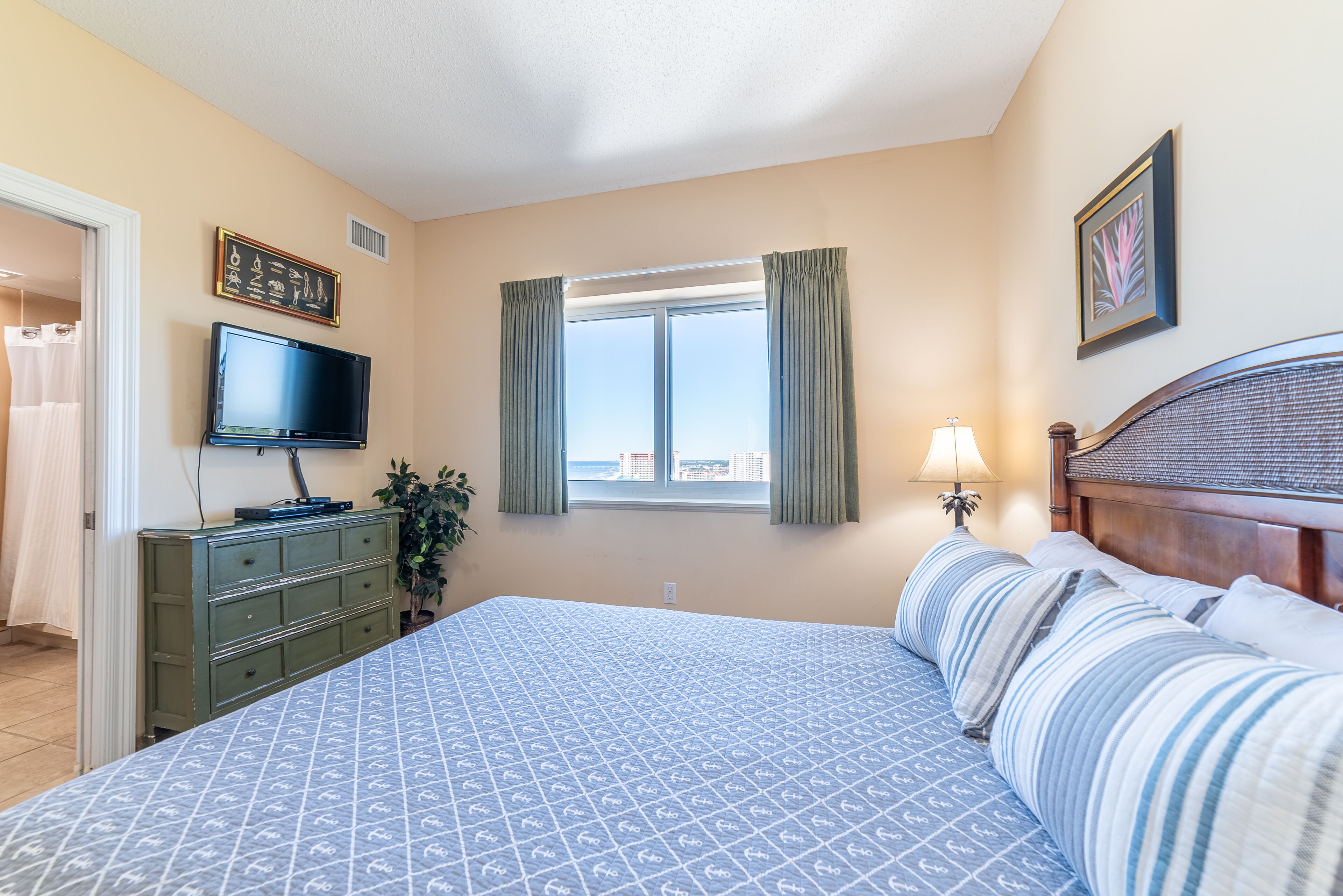 A bedroom with a bed, a TV, a dresser, a window with curtains, and a lamp.