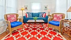 A living room with a blue sofa, two wicker chairs, a red and white patterned rug, and a ceiling fan. - Little House | Pet Friendly Cottage + Easy Walk to the Sand! (Tybee Island)