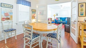 A dining area with a round wooden table and white chairs, a blue and white checkered window valance, and a "GONE TO THE BEACH" sign. - Little House | Pet Friendly Cottage + Easy Walk to the Sand! (Tybee Island)