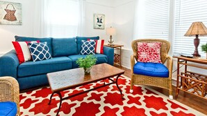 A living room with a blue sofa, a wicker chair, a patterned rug, and a small table with a lamp. - Little House | Pet Friendly Cottage + Easy Walk to the Sand! (Tybee Island)