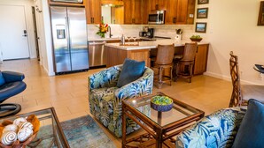 A modern kitchen with wooden cabinets, stainless steel appliances, and a central island. There is a dining area with a blue patterned sofa and a glass coffee table. - Ma'alaea Surf Unit E3-1 BR Ground Floor Beachfront Condo, Wi-Fi, AC, Pool, TV (Kihei)