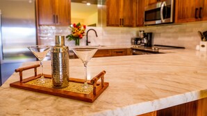 A kitchen with wooden cabinets, a stainless steel refrigerator, a microwave, and a marble countertop. - Ma'alaea Surf Unit E3-1 BR Ground Floor Beachfront Condo, Wi-Fi, AC, Pool, TV (Kihei)