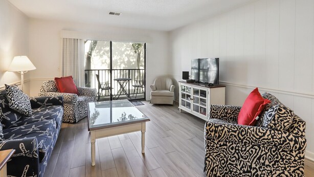 Ein Wohnzimmer mit gestreiften Sofas, einem Glastisch, einem Fernseher auf einer Kommode und einem Balkon mit einem Stuhl.