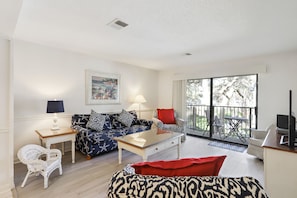 A living room with a sofa, coffee table, and a TV. There is a balcony with a table and chairs. - Surf Court 7 - Forest Beach Townhouse (Hilton Head Island)
