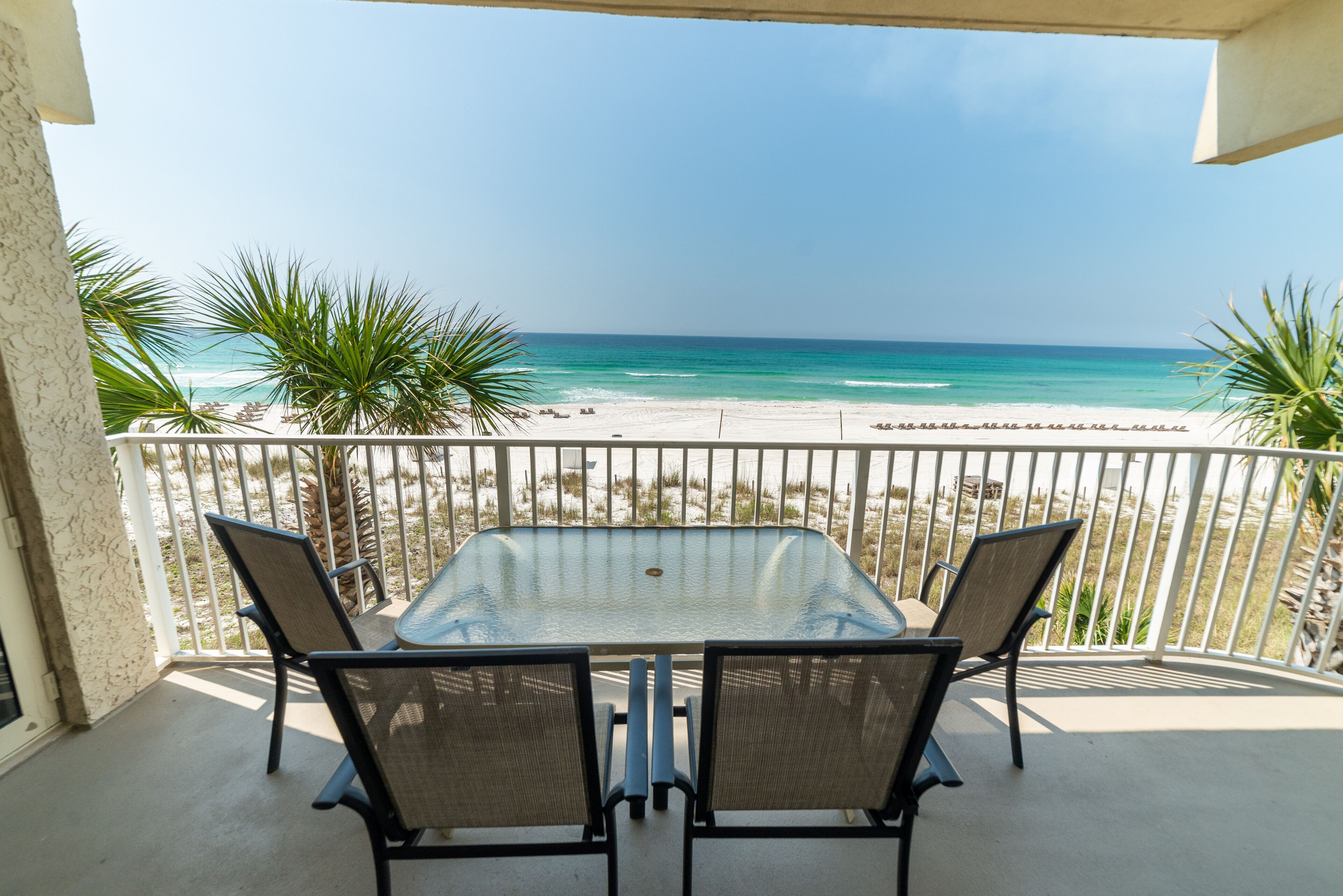 Een balkon met een glazen tafel en twee stoelen, uitkijkend op een strand met palmbomen en de oceaan.