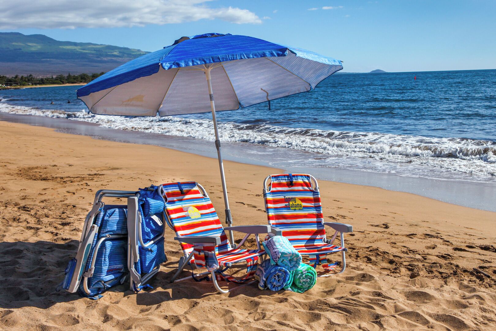 Una playa con dos sillas de playa a rayas, una sombrilla azul y una silla con una toalla.